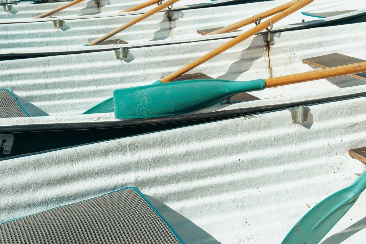 A close-up abstract shot of teal and wooden oars resting across the white hulls of rental boats, creating a bold pattern of intersecting lines and warm sunlight.